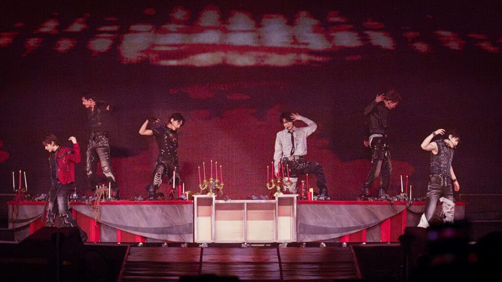 Six performers in dark outfits dance on a stage with red lighting, decorated with candles, drapes, and gold accents.