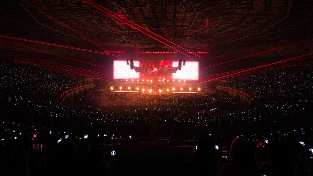 Crowded arena concert with red laser lights, large stage screens, and audience holding up glowing devices.