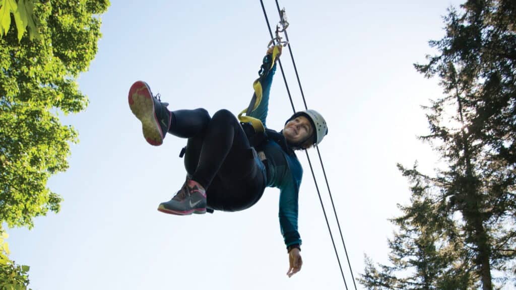 Person wearing a helmet and harness rides a zip line outdoors, surrounded by trees and a clear blue sky—a thrilling fusion of adventure and innovation inspired by the spirit of the PNW arts community.