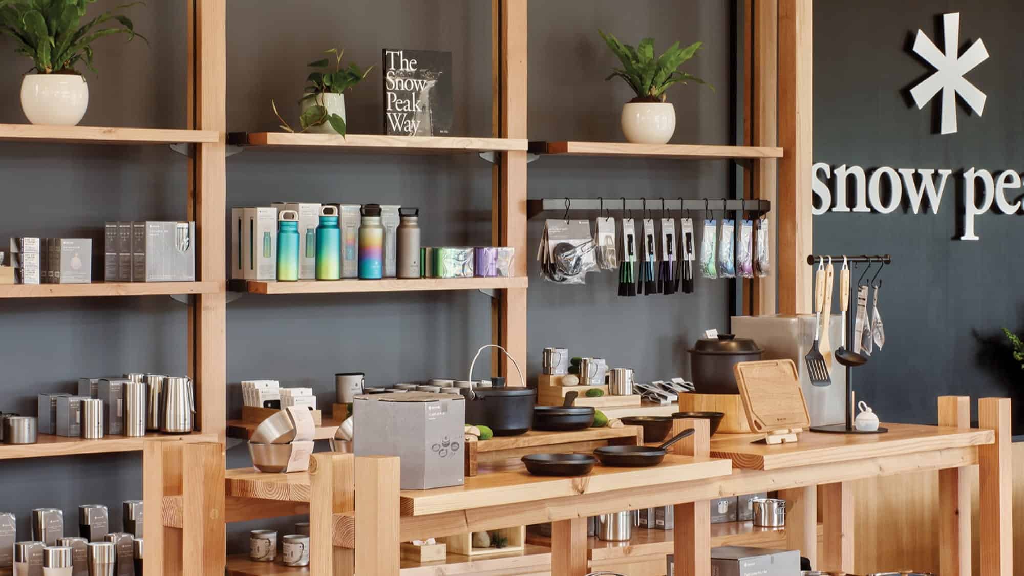 Wooden shelves display drinkware, kitchen items, and small accessories at a Snow Peak Outdoor Store, with potted plants and branded signage visible in the background.