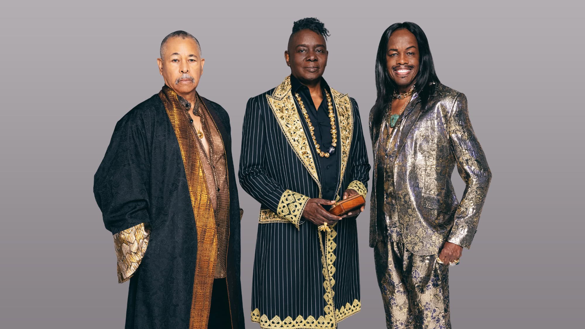 Three men stand side by side wearing ornate suits and coats with gold accents against a plain grey background.