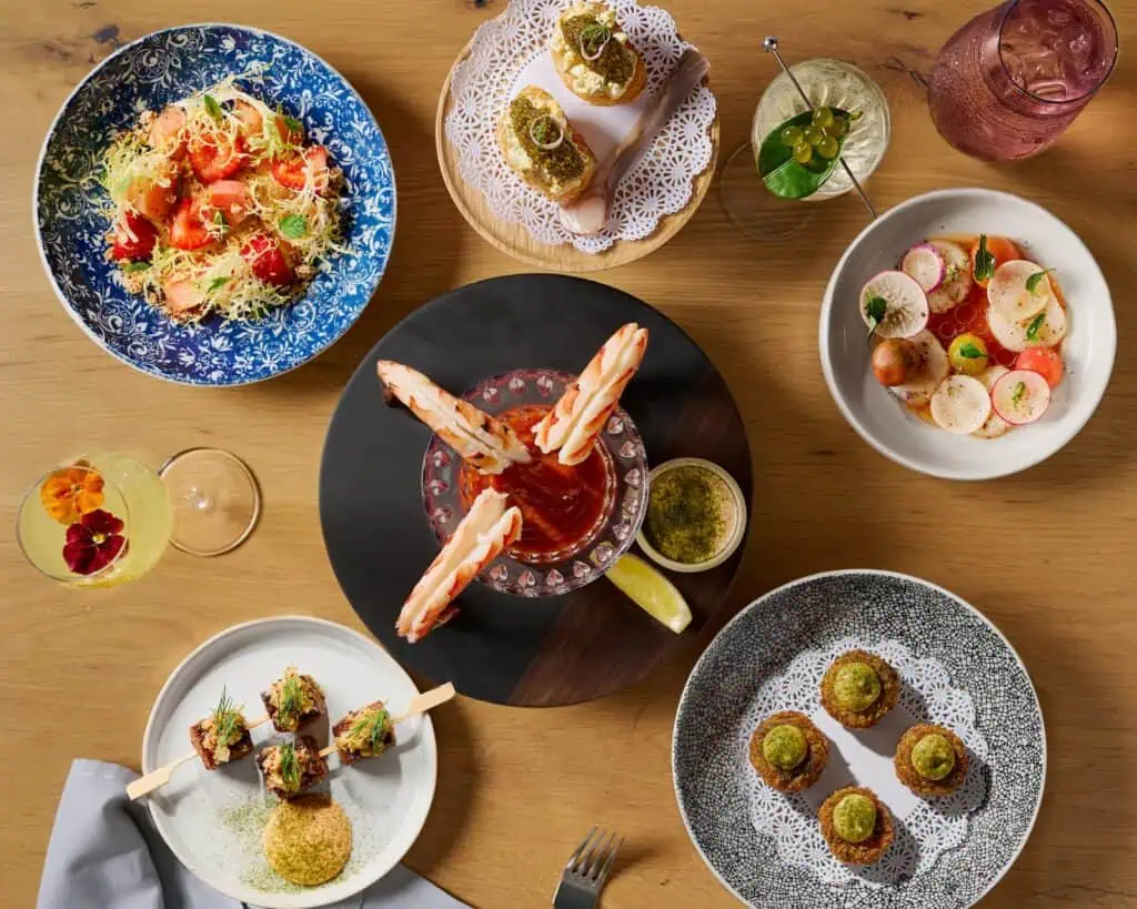 An overhead view of a wooden table set with assorted gourmet dishes, including shrimp cocktail, salad, skewers, and drinks.