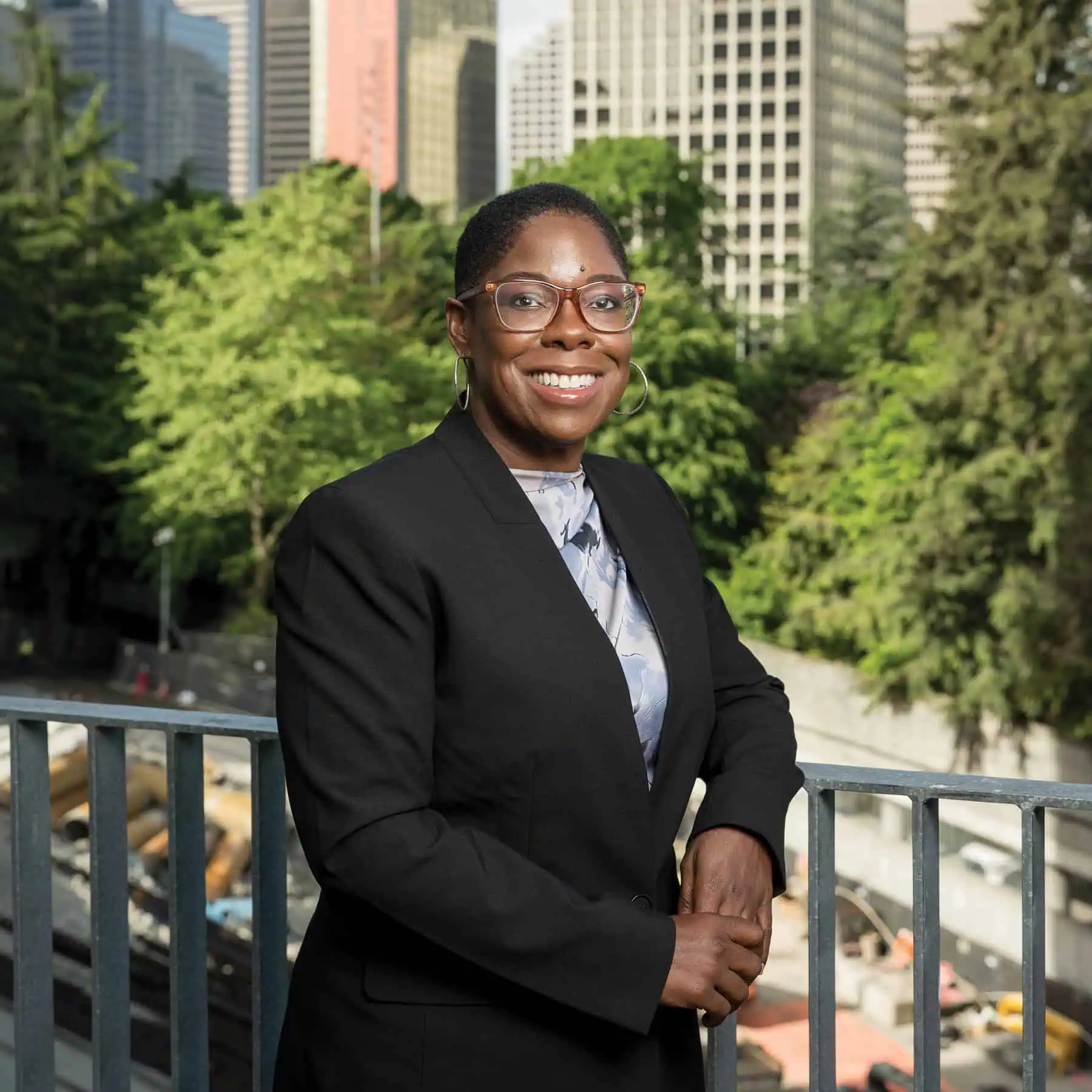A woman in a black blazer and glasses stands smiling on a balcony of a Senior living high-rise Seattle, with trees and tall city buildings in the background.