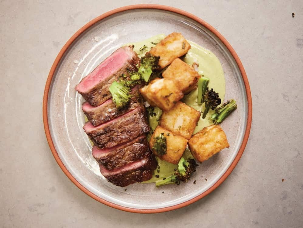 A plate with sliced medium-rare steak, crispy golden potato cubes, broccoli, and green sauce on a ceramic plate showcases the artistry of a renowned Seattle chef who brings Capitol Hill restaurant flair to every dish.