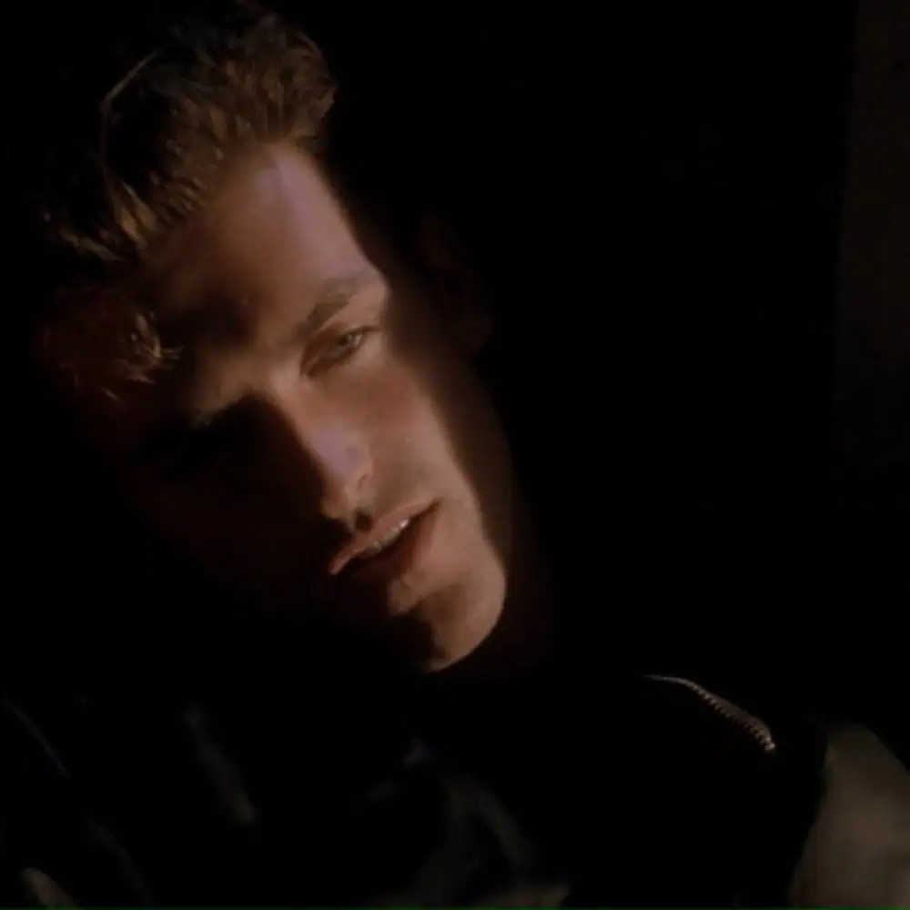A man with short hair is partially illuminated by a narrow beam of light, with most of his face in shadow, gazing slightly downward.