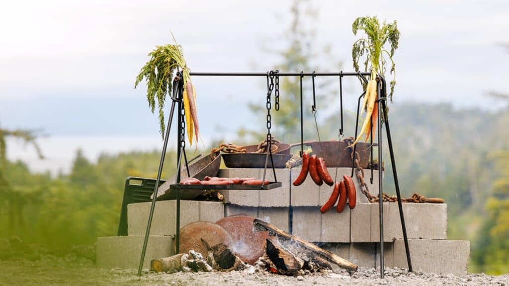 Outdoor grill with sausages, bacon, and vegetables cooking over an open fire, set up on cinder blocks with a natural landscape in the background.
