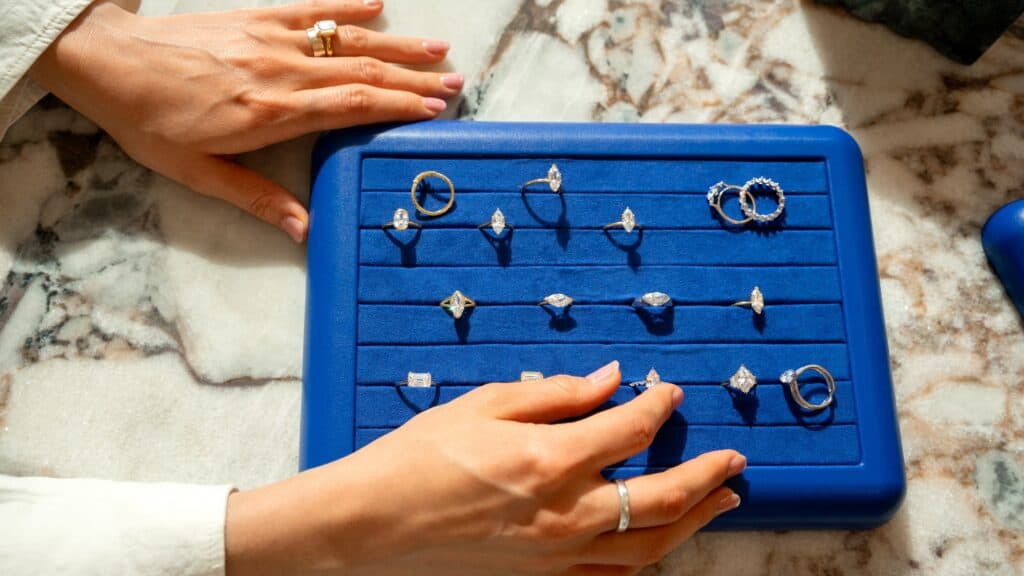 Hands near a blue display tray holding various gold and silver rings with gemstones, arranged in three rows on a marble surface.