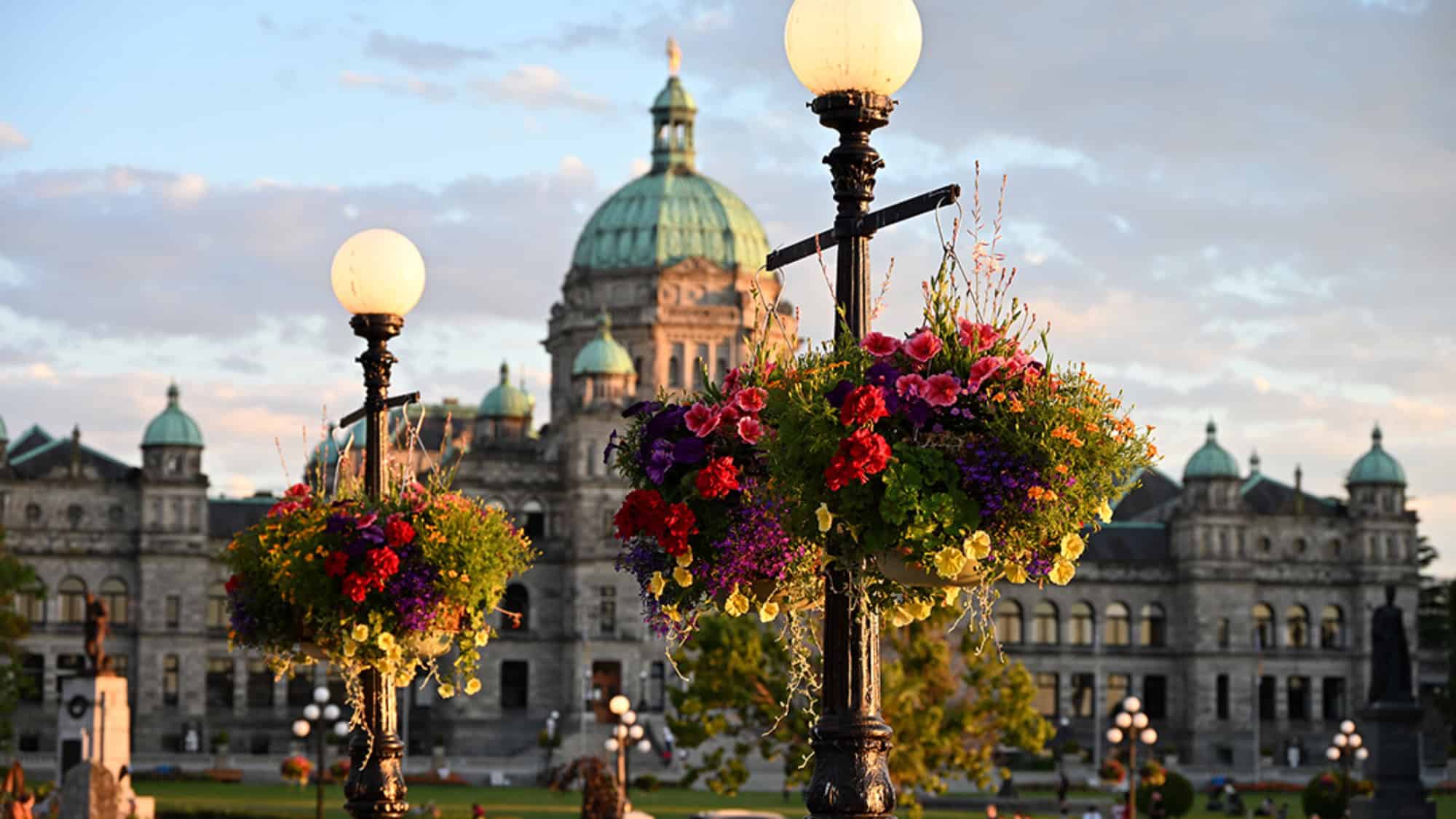 Hanging flower baskets on lamp posts in front of a domed historic building with green rooftops, under a partly cloudy sky.