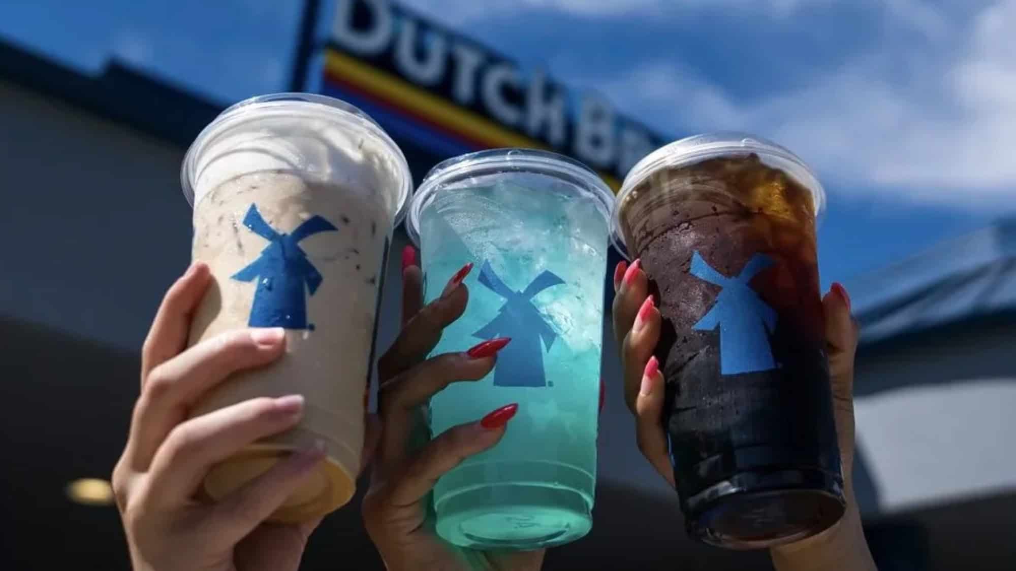 Three hands hold up large Dutch Bros cups—one with iced coffee, another with a striking blue beverage, and the third with an iced dark drink—in front of a Dutch Bros building sign. Make this stop part of your Restaurant Roundup for bold flavors and fun drinks.
