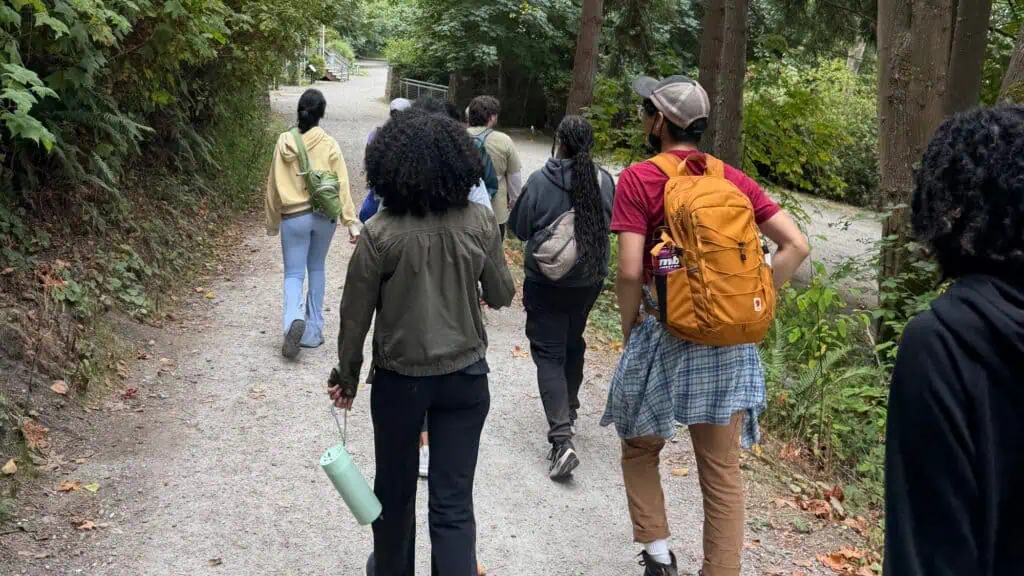 A group of people are walking on a gravel path through a wooded area, carrying backpacks and water bottles.