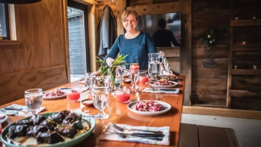 A woman stands smiling behind a wooden dining table set with plates of food, glasses, and candles in a cozy, rustic room.