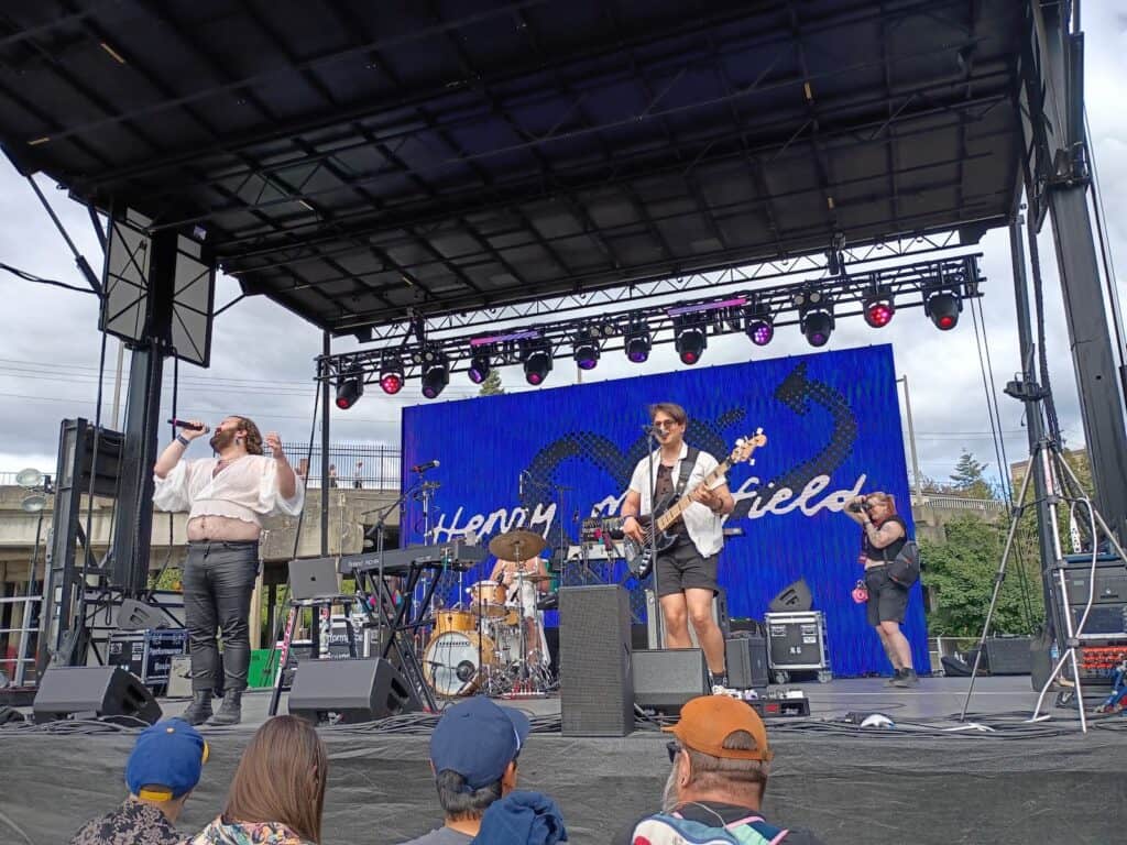 A band performs on an outdoor stage in front of a blue screen that reads "Henry Mansfield." Audience members watch from below on a partly cloudy day.