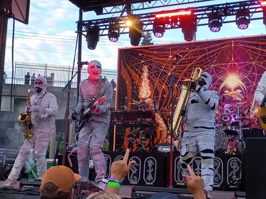 A band dressed as mummies performs on an outdoor stage with various instruments in front of a geometric patterned backdrop. Audience members watch and raise their hands.