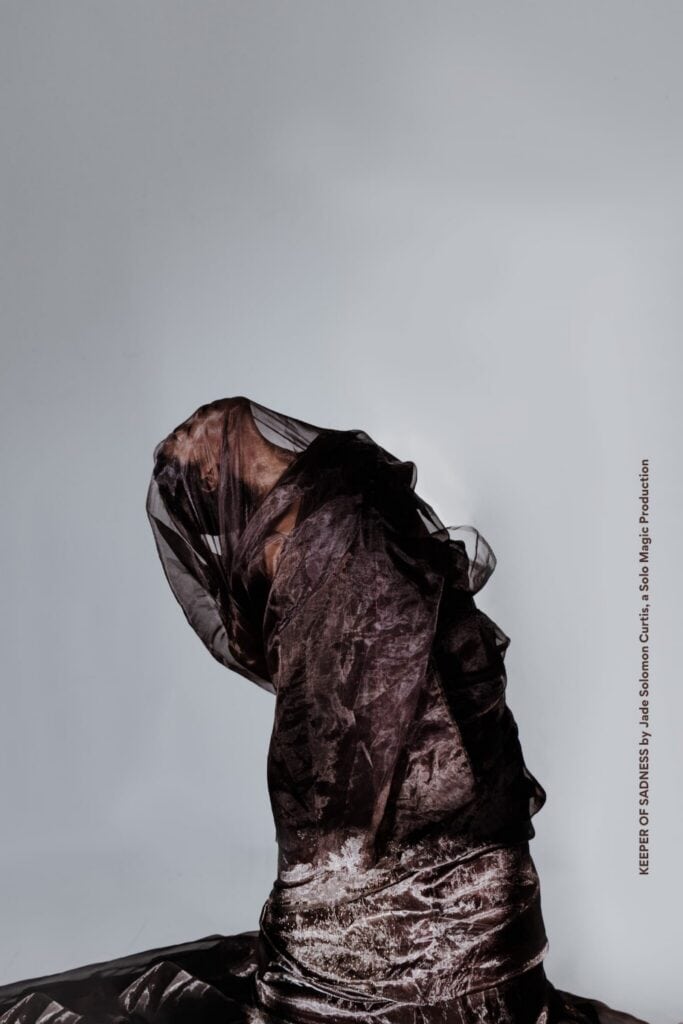 A person draped in sheer, dark fabric poses against a plain gray background, obscuring their face and body.