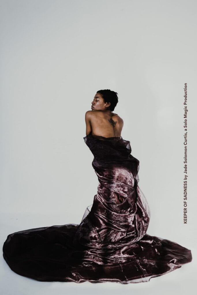 A person with short hair and a back tattoo stands wrapped in a flowing, dark velvet fabric against a plain white background. Text along the right edge reads "KEEPER OF SADNESS.