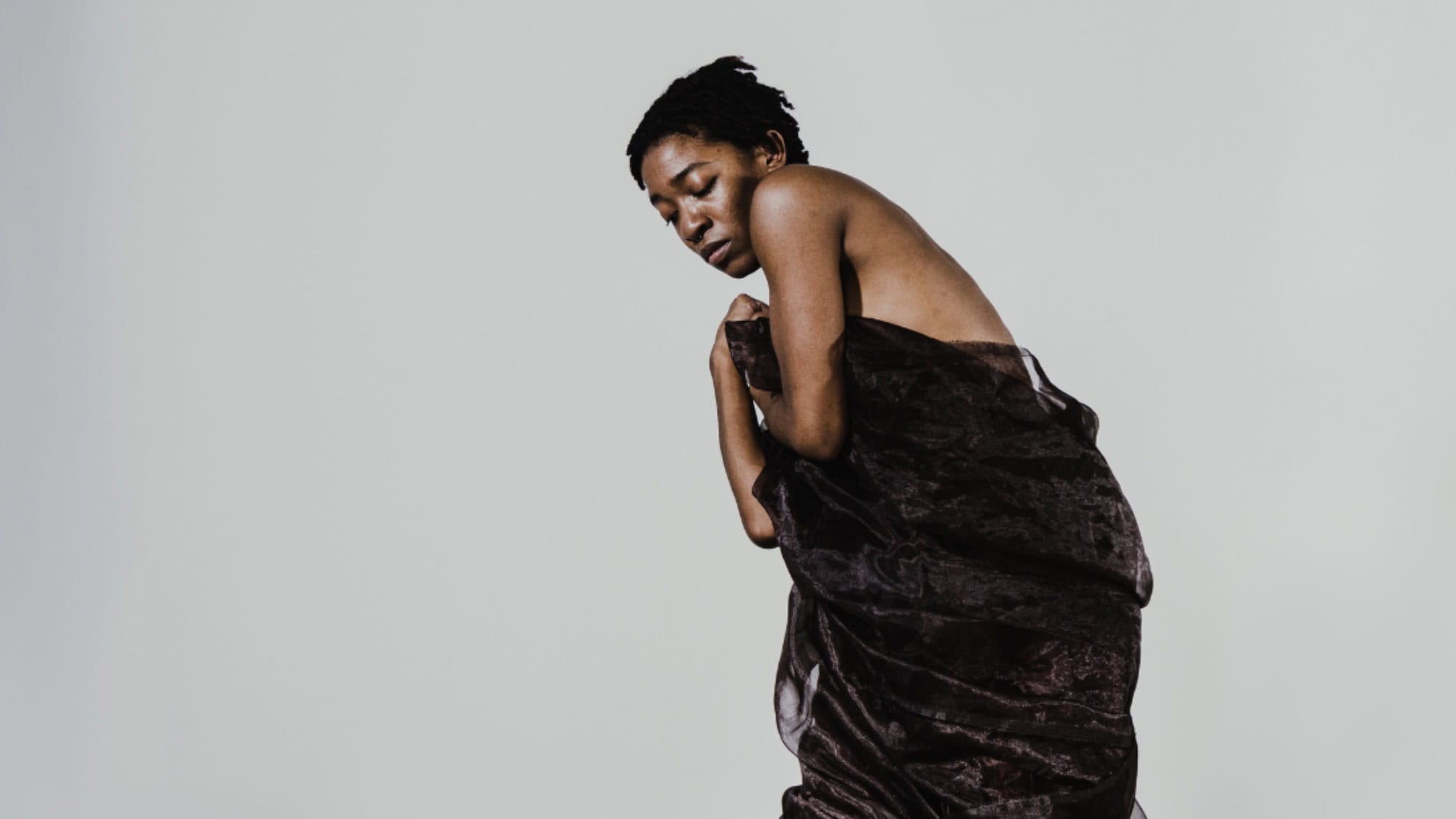 Person standing against a plain background, wrapped in a dark fabric, with eyes closed and head tilted slightly downward.