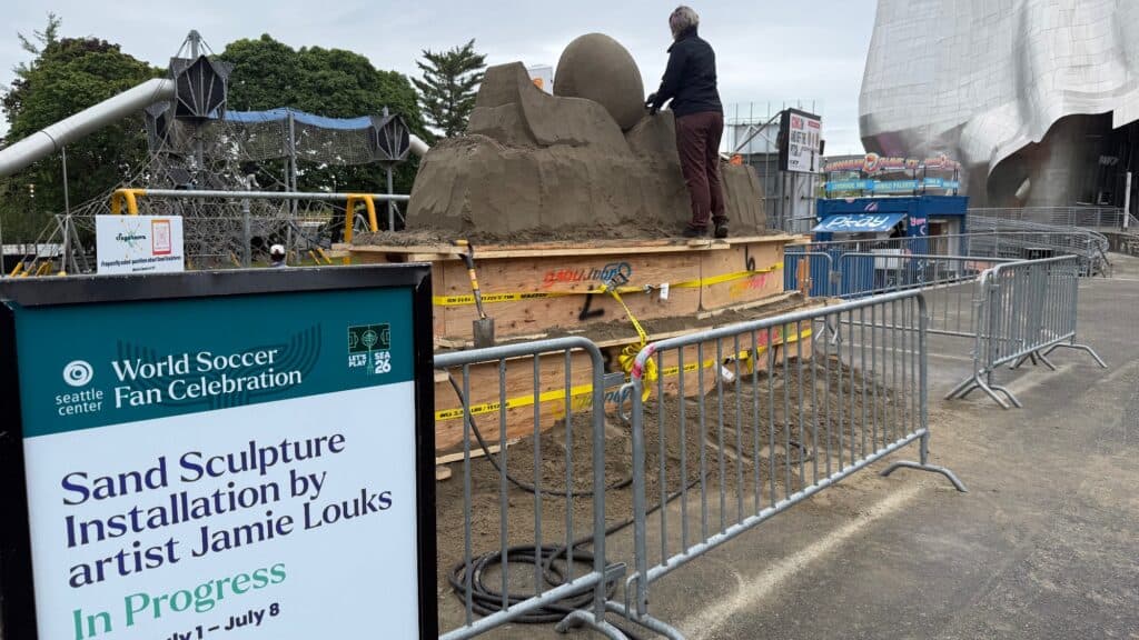 A person stands on a platform sculpting sand into a soccer-themed art installation. A sign reads "Sand Sculpture Installation by artist Jamie Louks. In Progress.