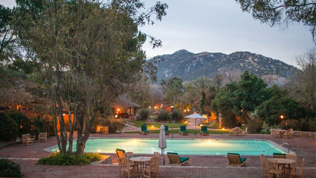 Rectangular outdoor pool surrounded by lounge chairs, tables, and trees, with mountains and greenery in the background at dusk.