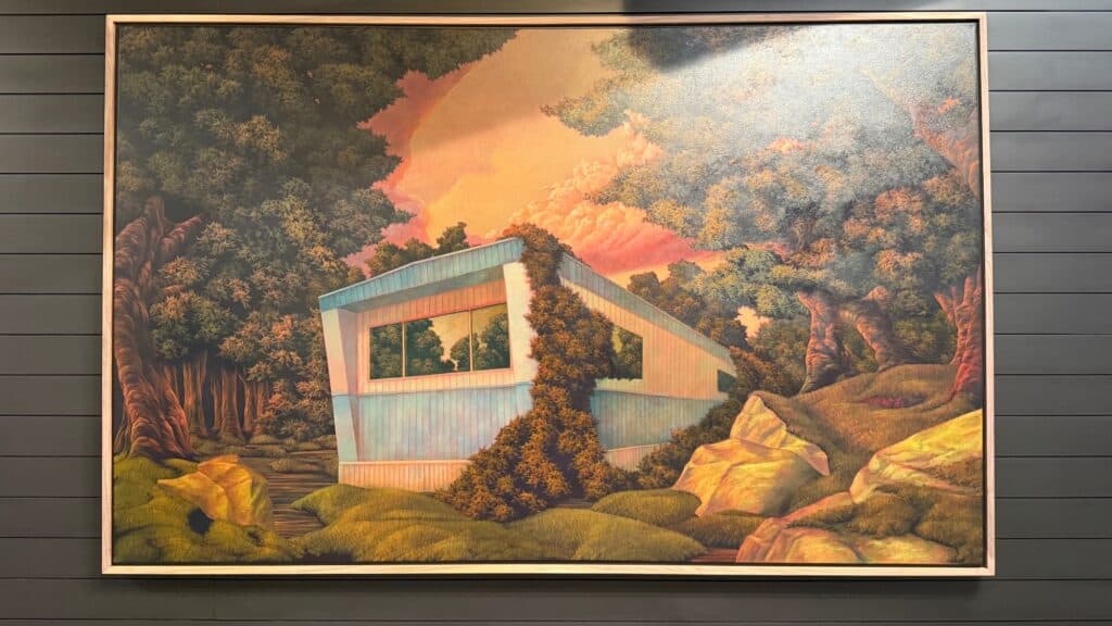 A framed painting of a blue mobile home in a forest surrounded by trees and rocks, with ivy growing on its side under a sky with orange clouds.