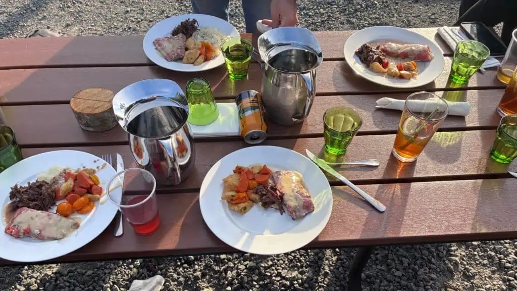 Four plates of food with meat, sauce, carrots, and potatoes on a wooden picnic table with drinks, glasses, metal pitchers, and utensils, outdoors on a gravel surface.