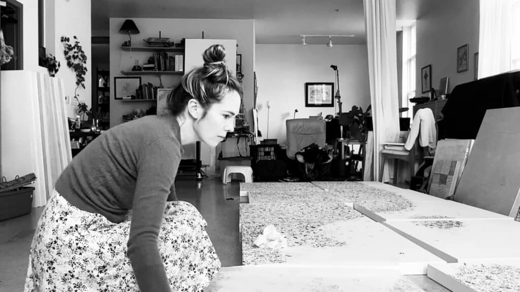 A woman kneels on the floor in an art studio, working on large canvases spread out in front of her.