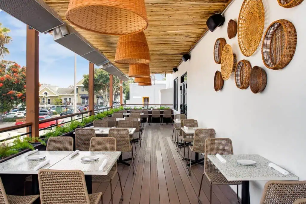 Outdoor restaurant patio with wicker chairs and tables, woven basket decor on a white wall, and large basket pendant lights hanging from a wooden ceiling.