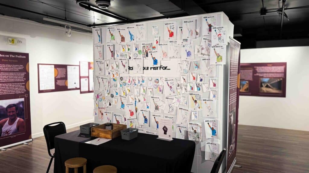 A display wall covered with colorful drawings behind a black table and two stools in a gallery setting evokes the creativity of a Summer Camp Hack, with informational panels on the surrounding walls.