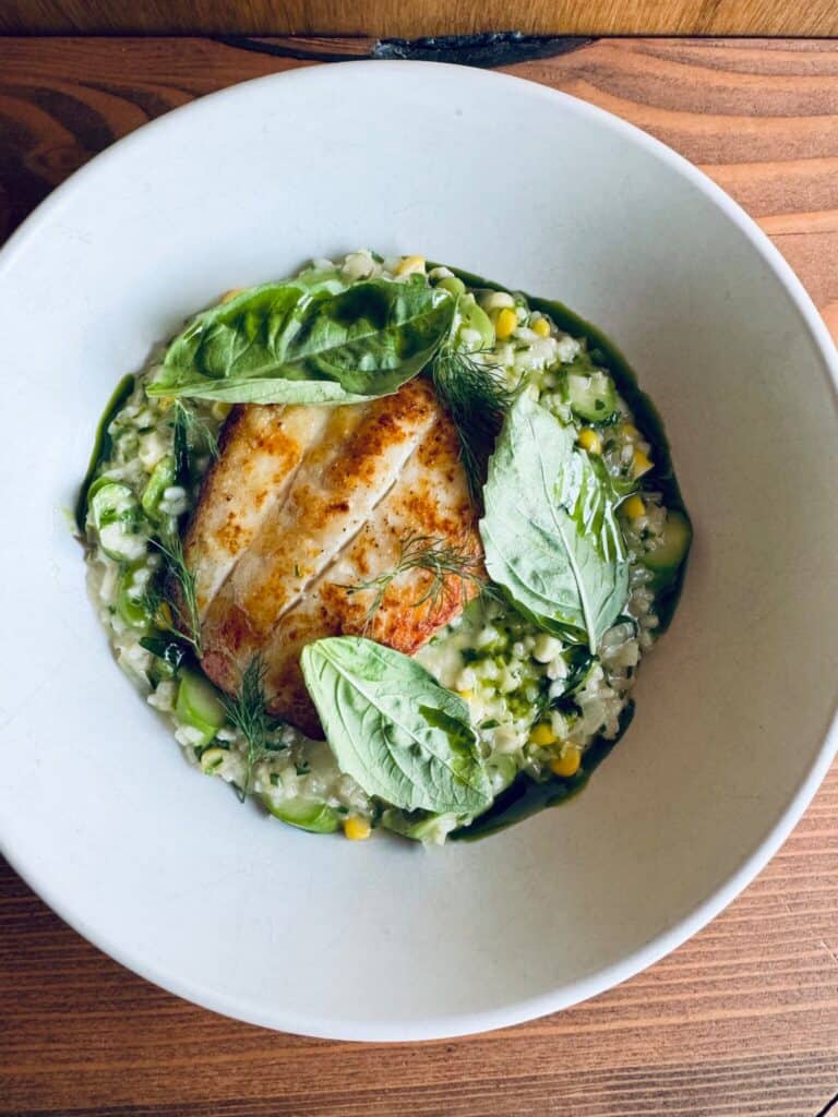 Grilled fish fillet on a bed of mixed vegetable risotto, garnished with fresh basil leaves, served in a white bowl on a wooden surface.