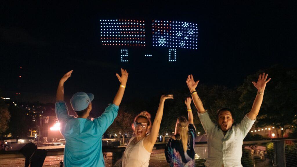 Four people cheering outdoors at night with two drone-formed flags and "2 - 0" displayed in the sky above them. Trees and city lights are visible in the background.