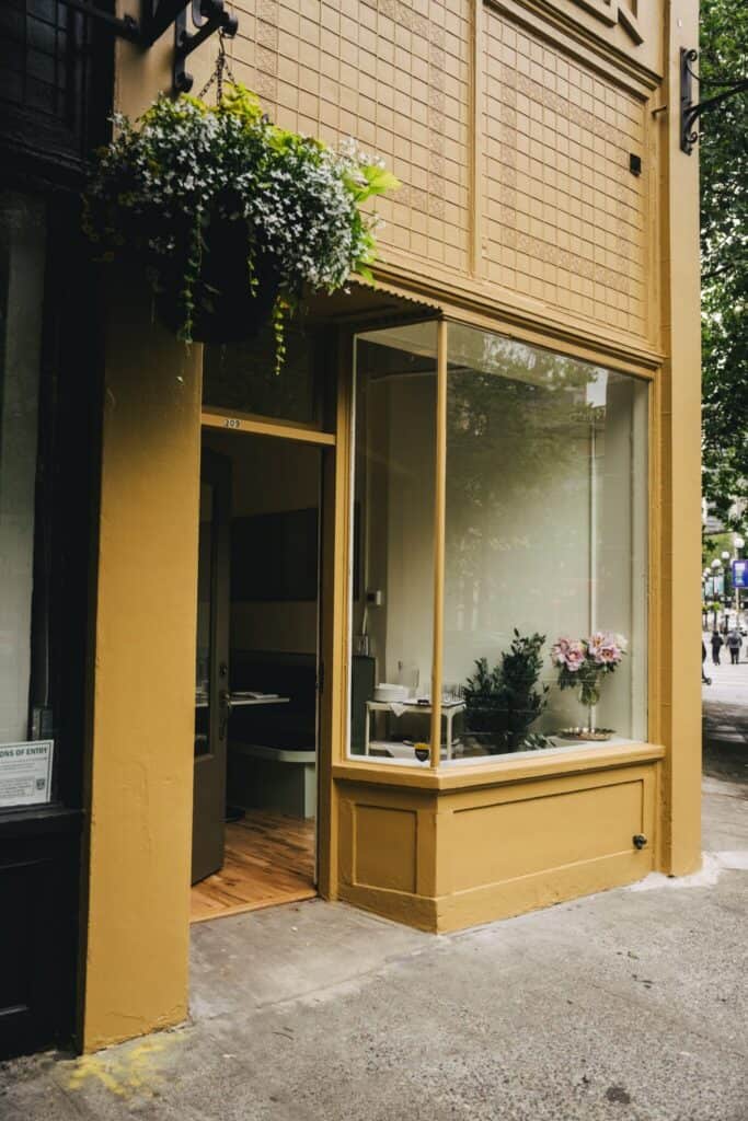 Storefront with large windows, yellow painted exterior, a hanging flower basket, and visible flowers and plants inside. The door is open, revealing part of the cozy restaurant interior—perfect for an inviting opening night in vibrant Pioneer Square.