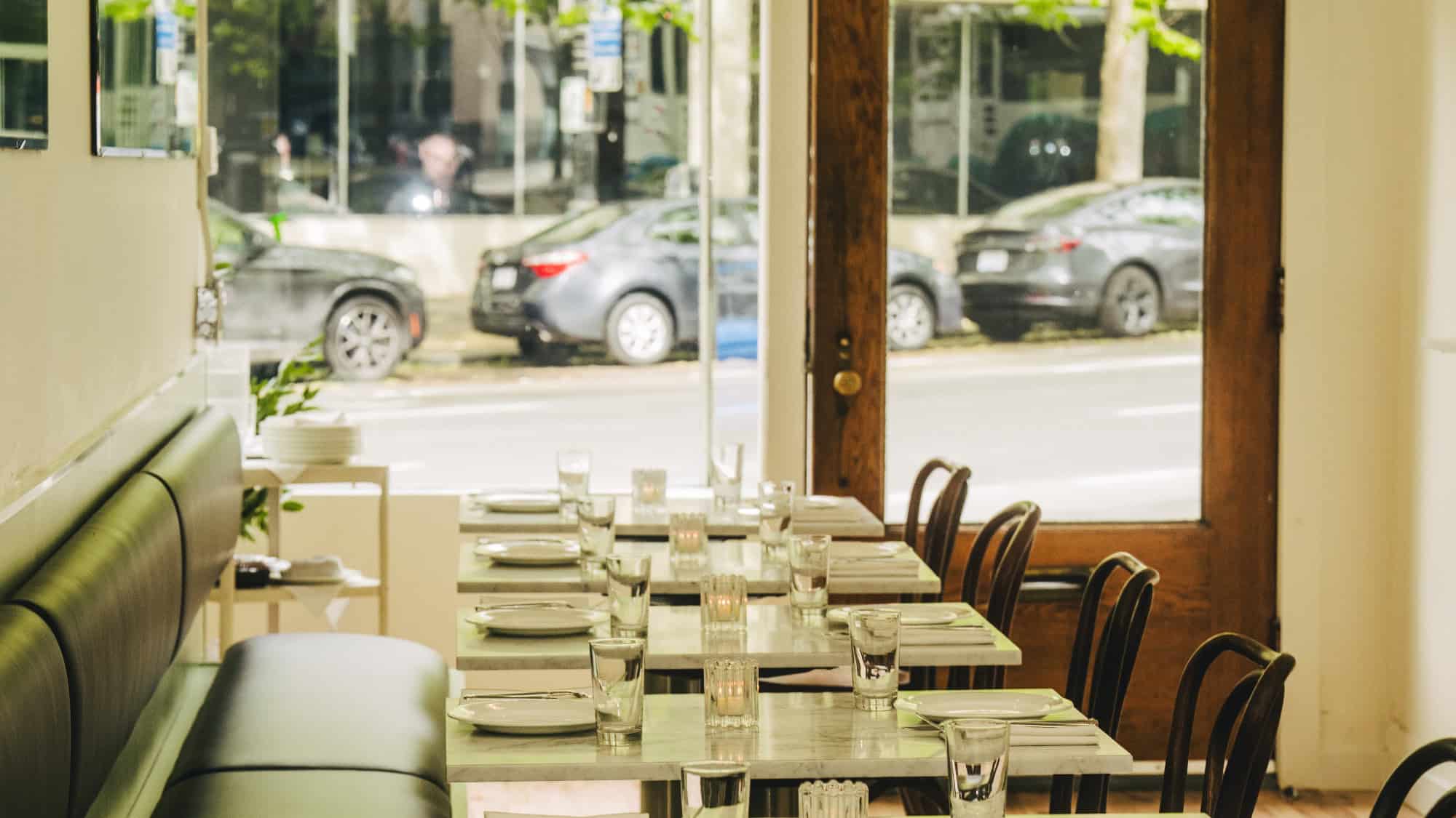 A set dining table with plates, glasses, and cutlery inside a restaurant, with large windows showing parked cars and trees outside.