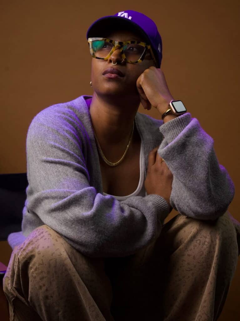 A person wearing a cap, glasses, sweater, and watch sits with their chin resting on their hand, looking upward against a brown background—a contemplative pose reminiscent of Jade Solomon Curtis during intimate Studio Sessions.