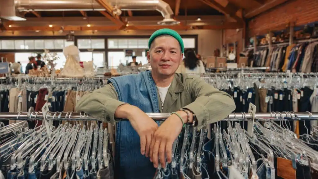 Person in a green hat leans on a clothing rack in a vintage clothing store, surrounded by various garments on hangers.