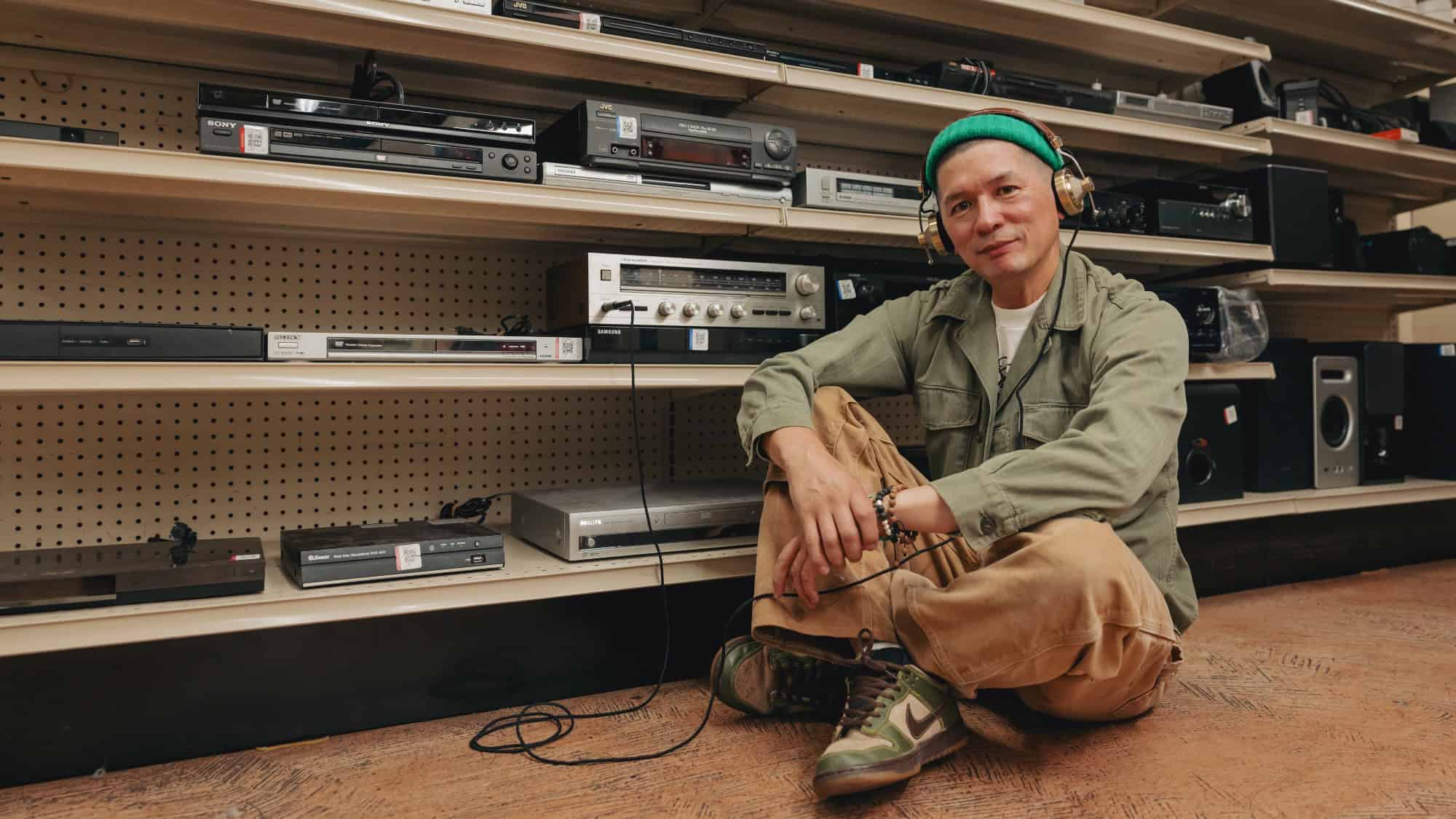 A person wearing a green beanie and headphones sits on the floor in front of shelves filled with vintage audio and video equipment.