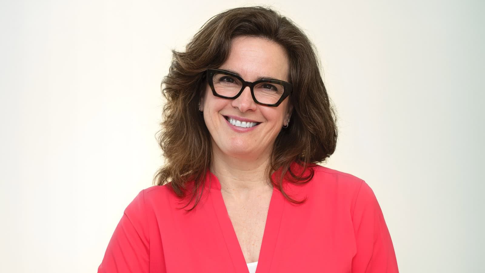 Woman with wavy brown hair and black glasses smiling, wearing a bright pink blouse, standing in front of a plain light background—her warm presence reflects her passion for longevity and family medicine.