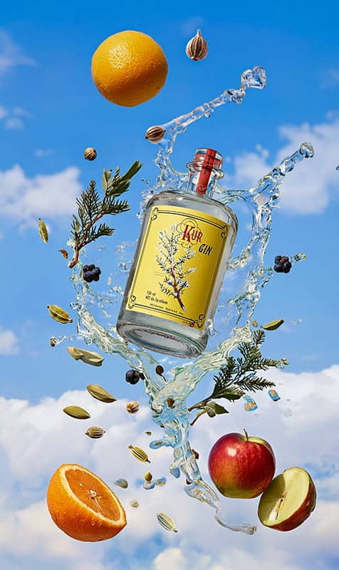 A bottle of gin by Wildwood Spirits Co, a renowned PNW spirits craft distillery, is surrounded by splashing water, orange slices, apple, spices, and botanicals against a blue sky with clouds.