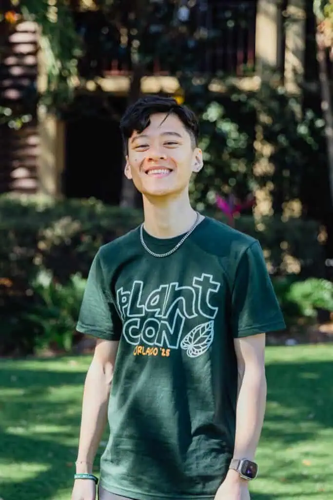 A person stands outdoors on grass, smiling, wearing a dark green “PlantCon Orlando ’23” T-shirt and a watch, with trees and plants in the background—bringing a touch of Seattle energy to PlantCon as if ready to fill windowsills everywhere with greenery.