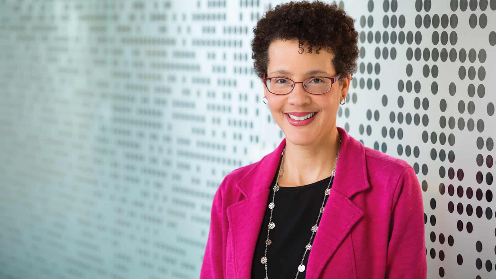 Dr. Kristin Conn, a dedicated family medicine physician at Kaiser Permanente, stands in front of a patterned wall featuring rows of black dots. She wears a pink blazer and necklace, her short curly hair and glasses complementing her professional yet approachable demeanor.