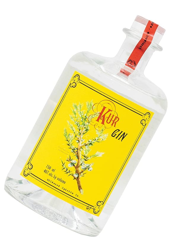 A clear glass bottle of Ikuer Gin by Wildwood Spirits Co, with a yellow label featuring a juniper branch illustration and a red seal on the neck—an award-winning, farm-to-bottle spirit.