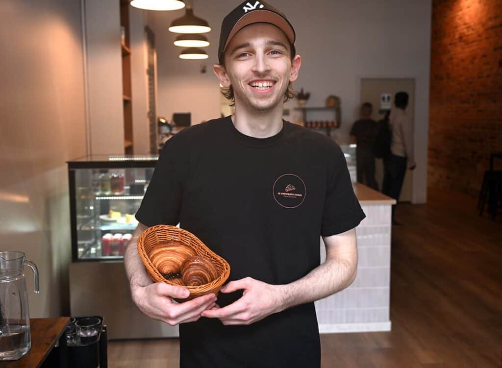 A person wearing a black t-shirt and cap smiles while holding a wicker basket of pastries inside a cafe with a counter and display case in the background.