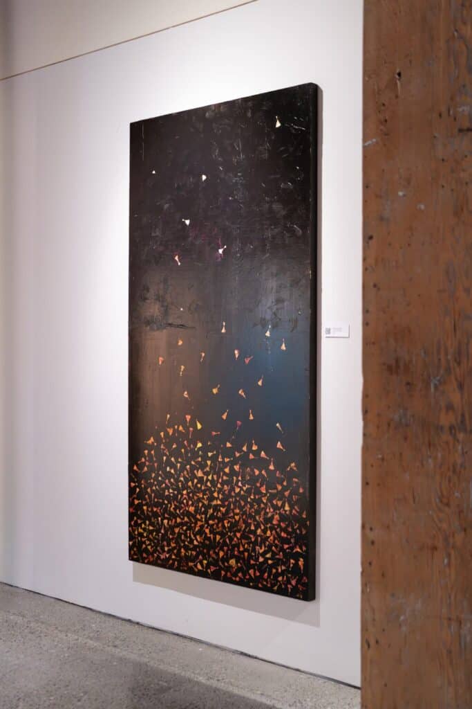 Large vertical painting with a black background and clusters of small, colorful triangles concentrated at the bottom, gradually dispersing upward. Displayed on a white gallery wall.