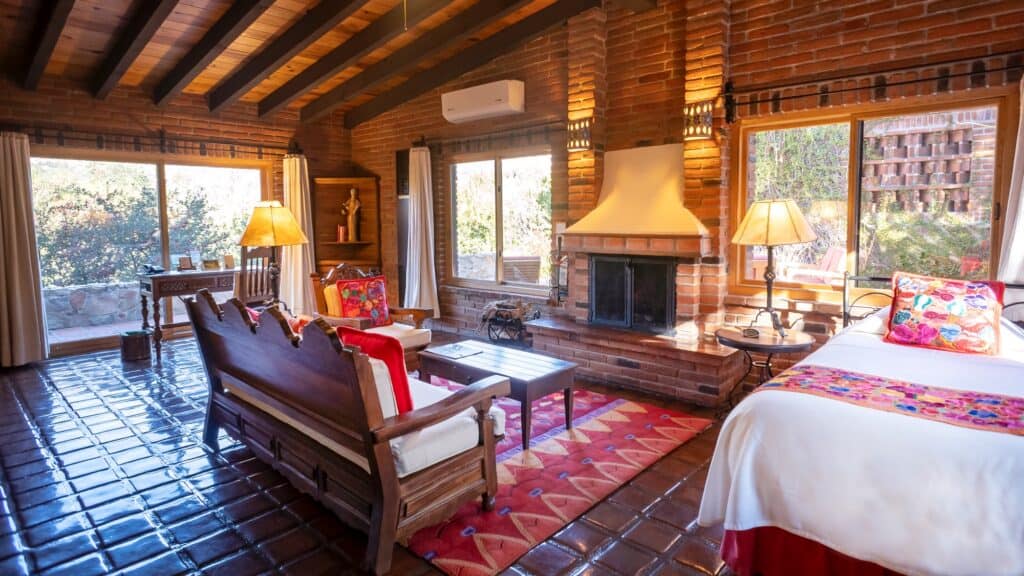 Cozy room with brick walls, large windows, a fireplace, wooden ceiling beams, tiled floor, and rustic furniture, including a bed, chairs, and a table with colorful cushions and lamps.