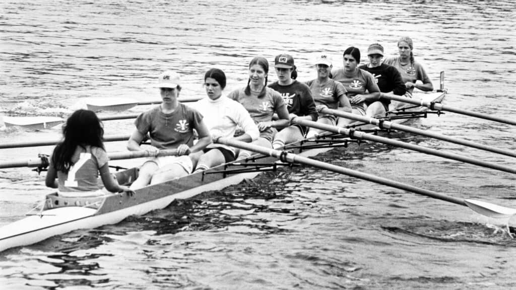 Eight rowers and a coxswain in a racing shell on the water, all wearing matching shirts and rowing in sync.