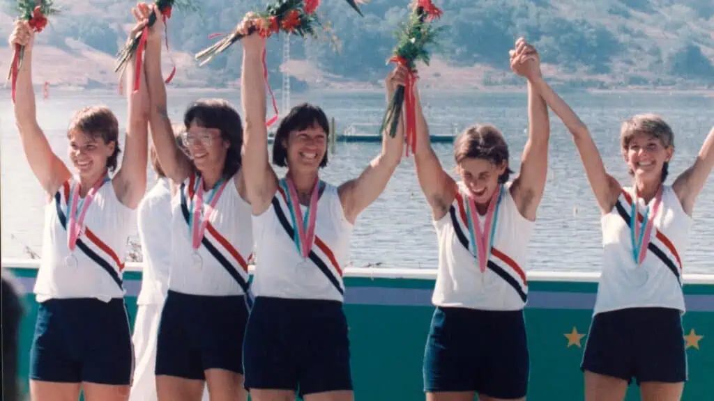 Five female athletes in matching uniforms stand on a podium, holding flowers and medals, with their arms raised in celebration by a lakeside.