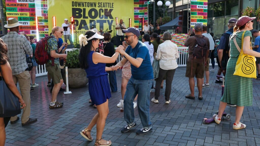 People dance and socialize at the Downtown Seattle Block Party, with a bright event banner and colorful decorations in the background.