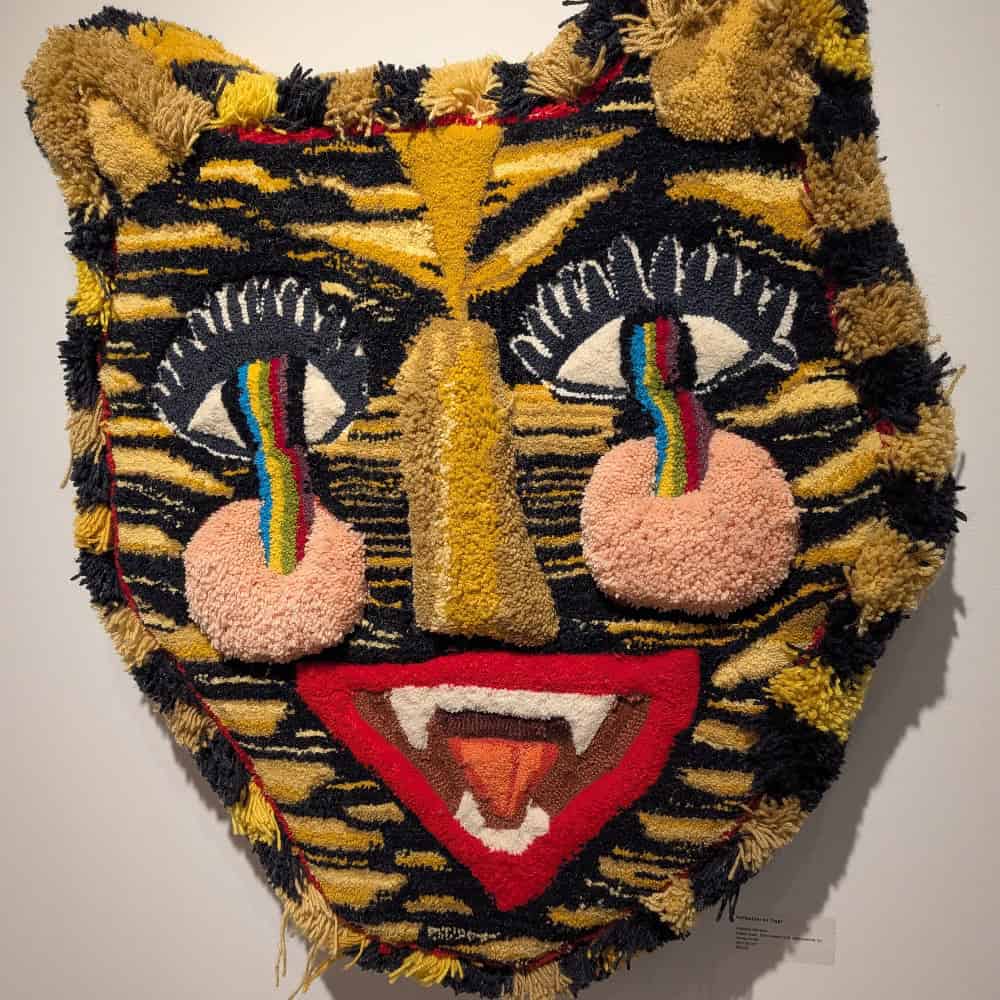 Textile art of a cat-like face with yellow and black stripes, large red mouth with fangs, pink cheeks, and rainbow tears under white eyes.