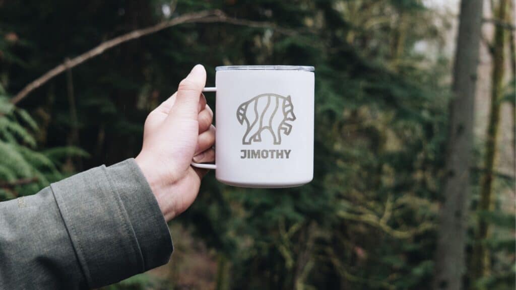 A hand holds a white metal mug with a bear graphic and the name “Jimothy” against a blurred forest background.