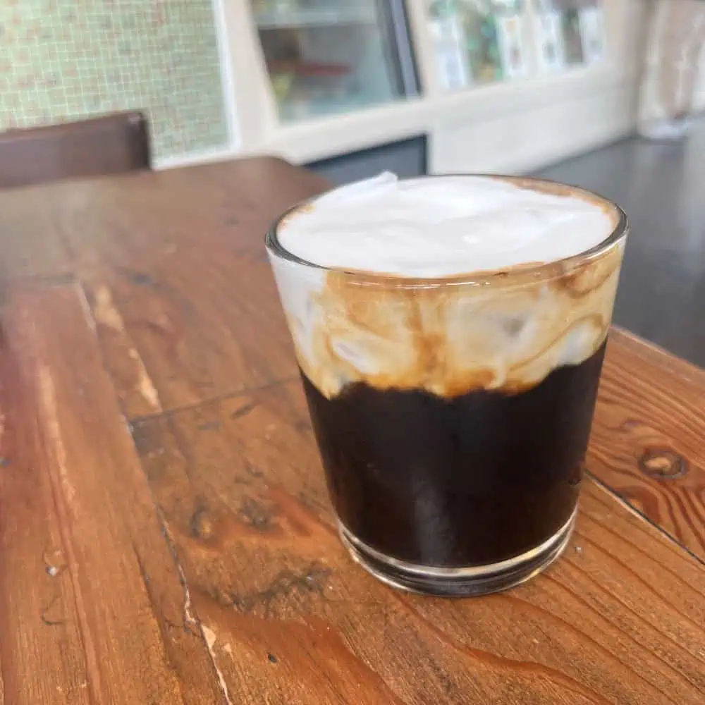 A glass of iced coffee topped with a layer of foamed milk sits on a wooden table in a cafe setting.
