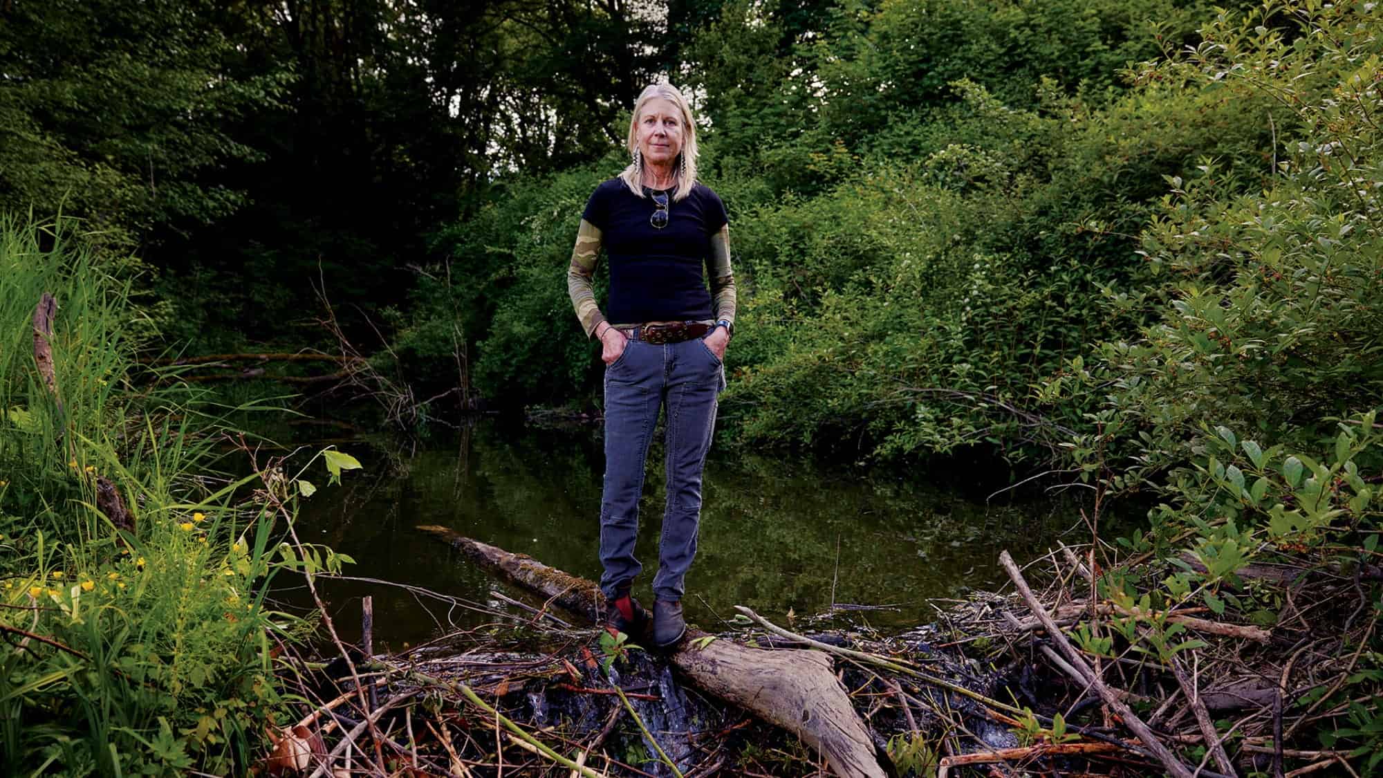 A person with long gray hair stands on a log by a pond surrounded by dense green foliage, wearing a black shirt and gray pants, clearly having done a Dam Good Job finding the perfect spot to enjoy nature.