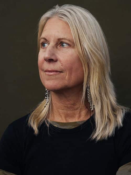 A woman with long blonde hair and feather earrings looks slightly to the side against a plain dark background, exuding quiet confidence as if she just finished a Dam Good Job.
